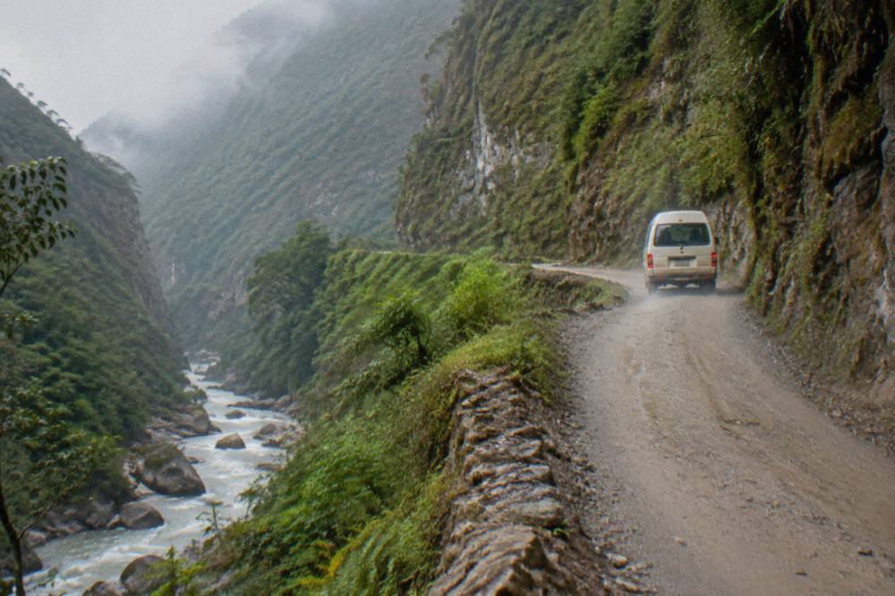 Narrow dirt road beside a river gorge with green cliffs and a small van rounding a bend