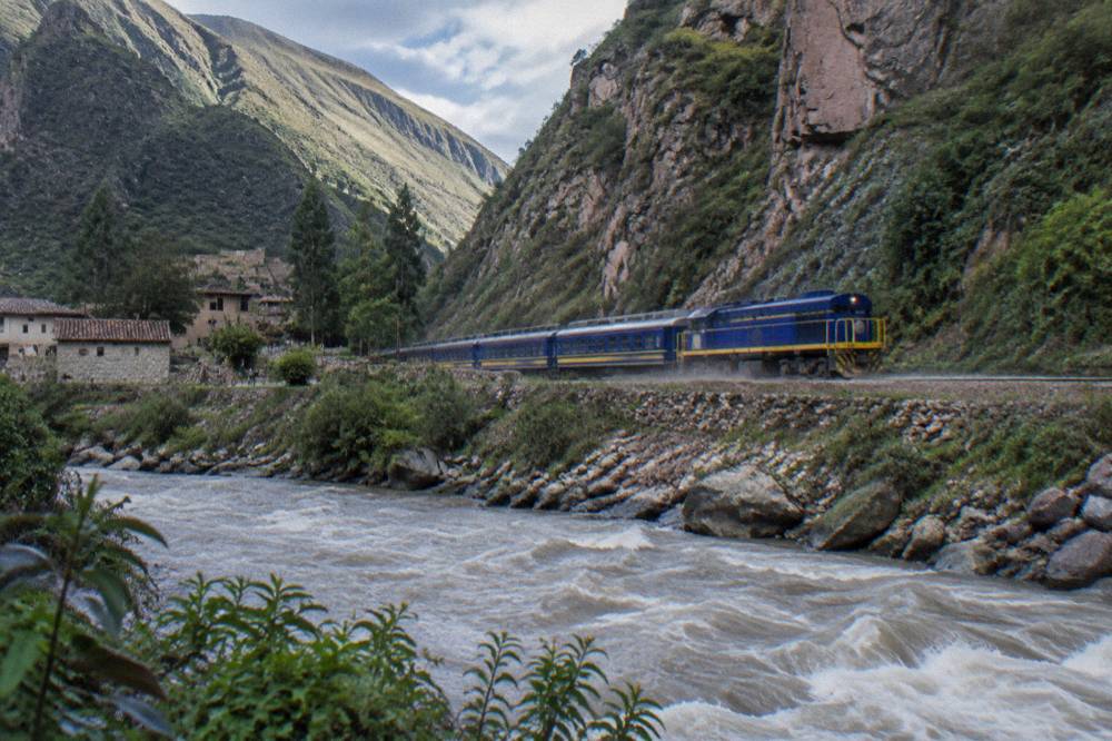 Blue and yellow passenger train on rails beside a rushing river between steep green canyon walls