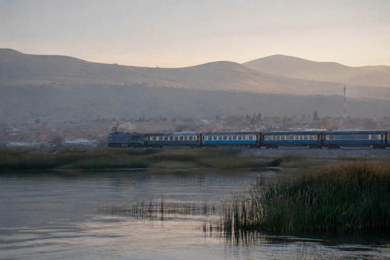 A passenger train traveling along the shoreline of Lake Titicaca with high Andean hills behind it