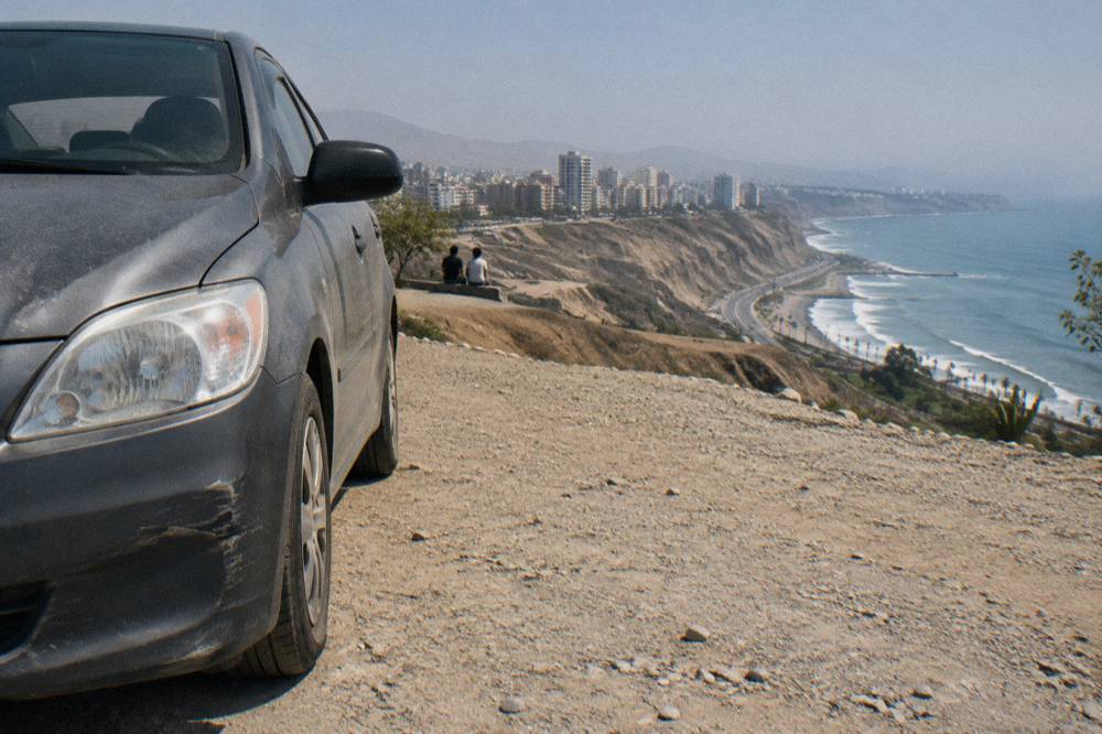 A rental car parked at a viewpoint above a desert road stretching toward coastal cliffs