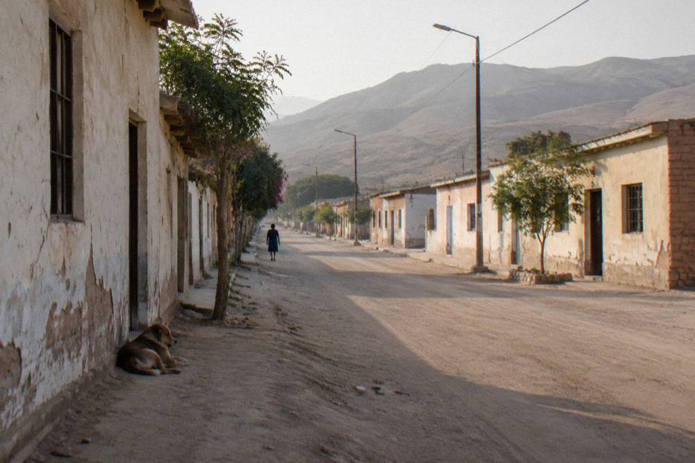 Desert town street with low buildings and dry hills rising under a hazy afternoon sky