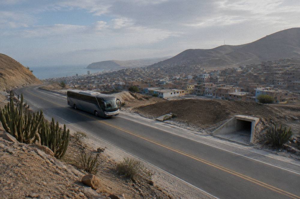 A modern touring coach traveling on a paved two-lane highway through arid hills in southern Peru