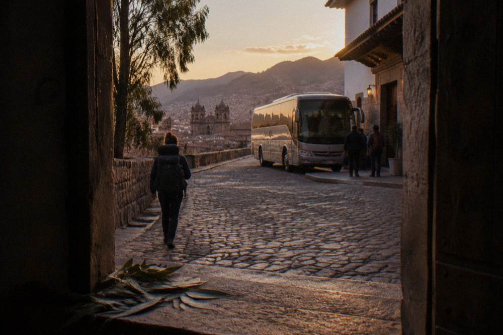 A traveler with a backpack watching a tour bus arrive at a hotel entrance on a quiet city street in the evening