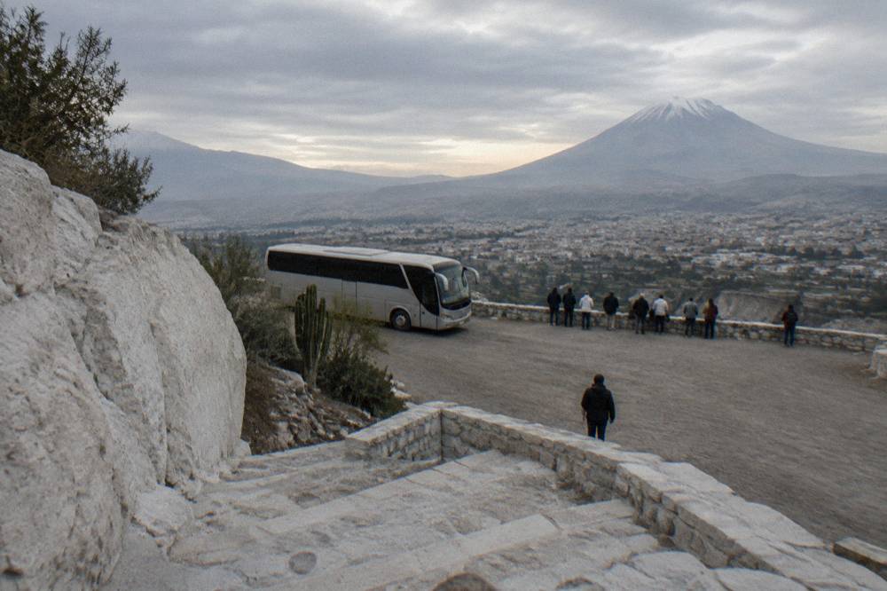 A tour bus parked at a high mountain pass with travelers standing at a viewpoint