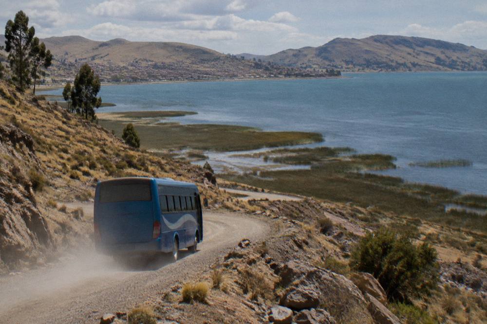 Blue tourist bus on a mountain road above the brown shoreline of Lake Titicaca under a wide sky