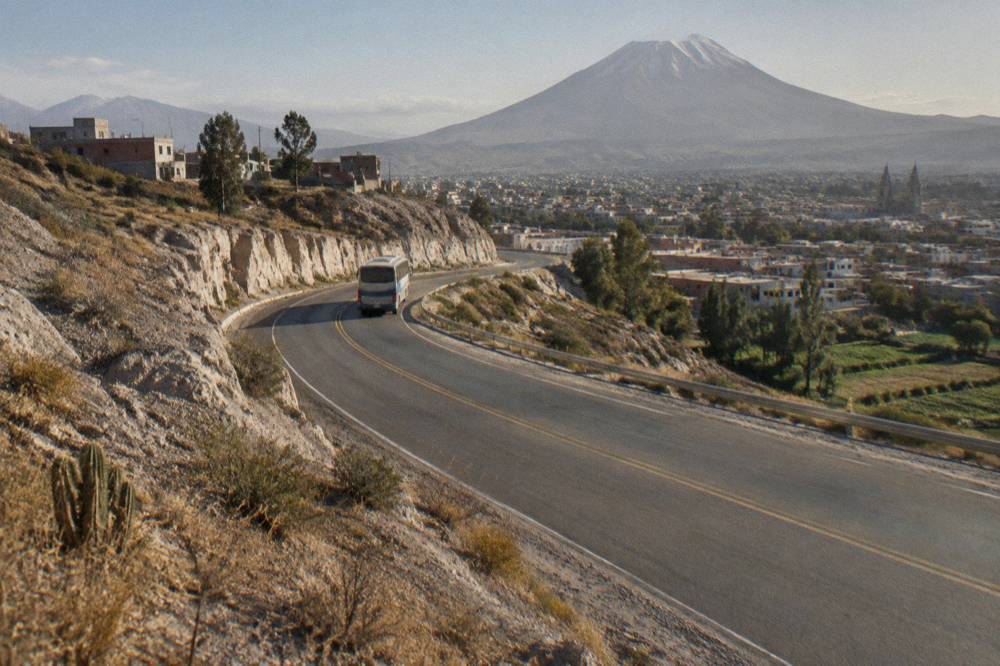 Long-distance bus on a paved highway curving between dry hills in late afternoon light