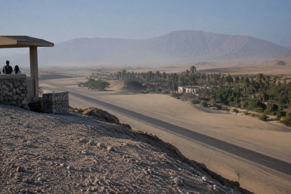 A desert highway crossing pale sand dunes with an oasis of palm trees visible in the distance
