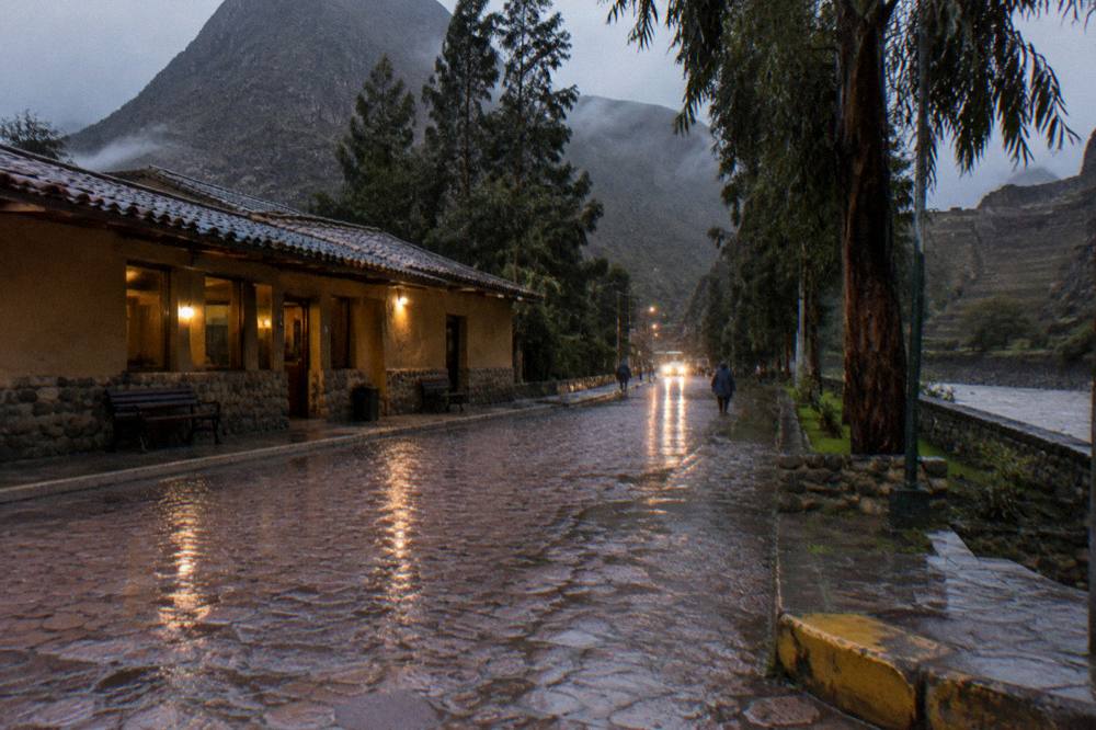 An evening street outside a bus terminal with a coach's headlights on and wet pavement reflecting the lights