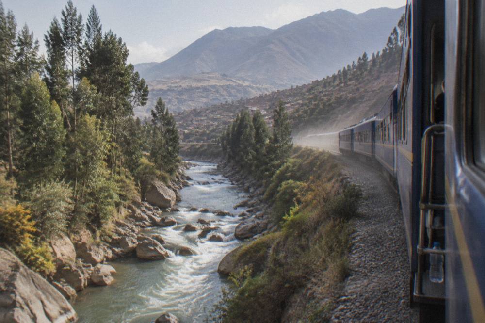 A scenic tourist train travelling along a river gorge toward green mountains