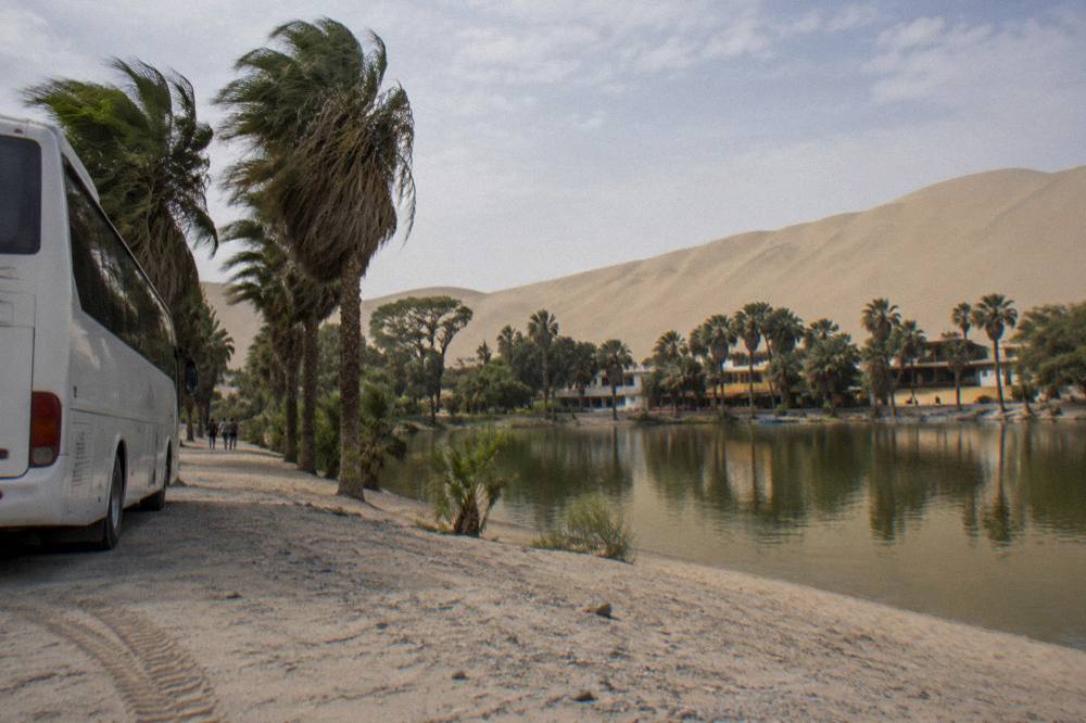 A tour bus parked beside the Huacachina oasis lagoon with sand dunes rising behind it