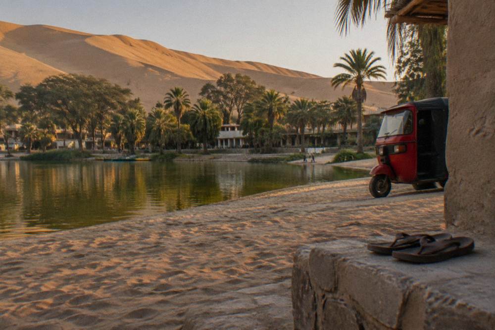 A desert oasis village with a lagoon ringed by palm trees and tall sand dunes under a clear sky