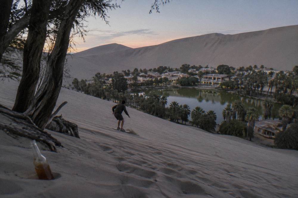 A sandboarder descending a tall dune above the Huacachina oasis lagoon at golden hour