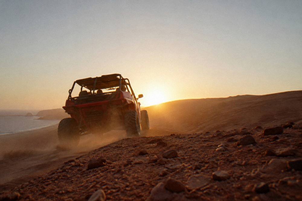A dune buggy cresting a steep sand slope with a low sun behind it