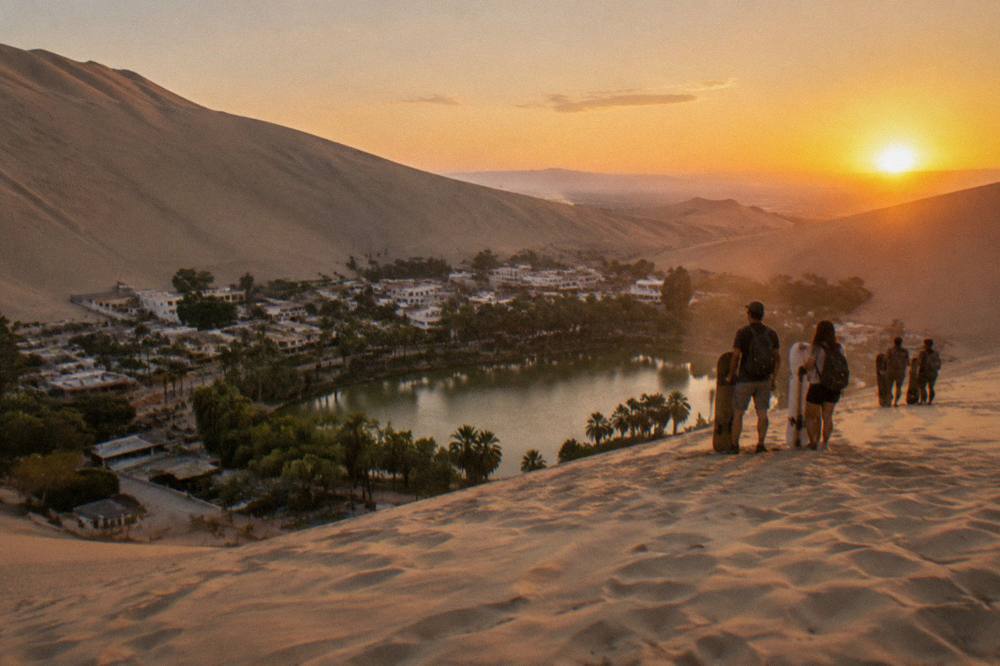Group of travelers with sandboards standing on a tall dune at sunset