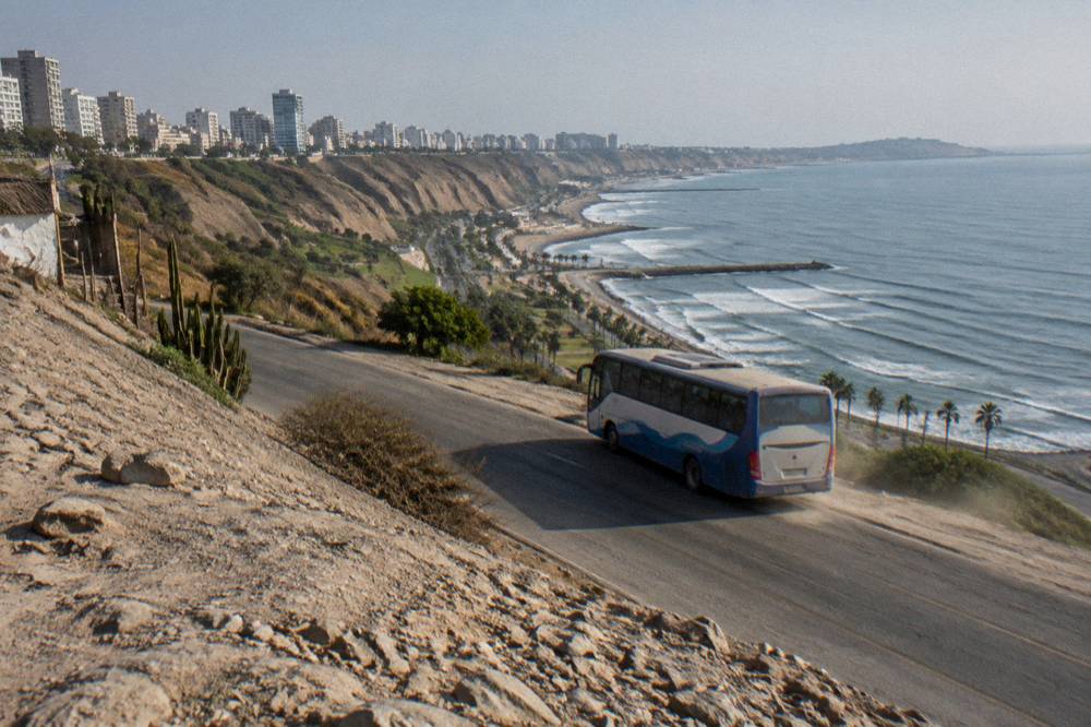 A touring bus winding along a road above a high Andean lake with snow-capped peaks beyond