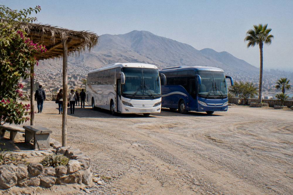 Two buses parked side by side at a highway rest stop with mountains behind them
